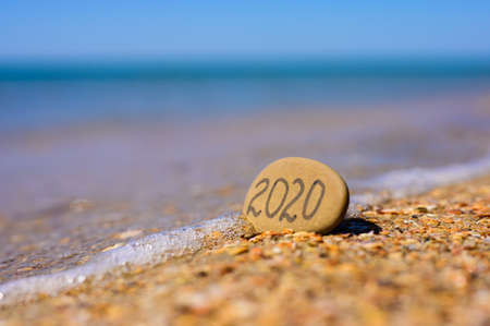 Sea water washes the pebbles with the inscription 2020 on the beach. Sea vacation symbol in 2020の写真素材