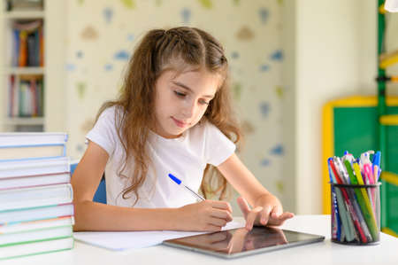 The girl does homework and uses the tablet. Girl schoolgirl sitting at a desk at homeの写真素材