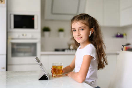 The girl sits at home at the table and holds a glass of juice in her hand. Nearby is a tablet. girl looking at the camera and smiling cuteの写真素材