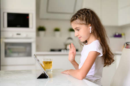 The girl positively communicates on the Internet with friends. Girl at home sitting at a white table with a tablet. Nearby is a glass of juice.の写真素材