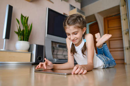 A girl in a white T-shirt lies on the floor. Girl playing on a tablet and smiling.の写真素材