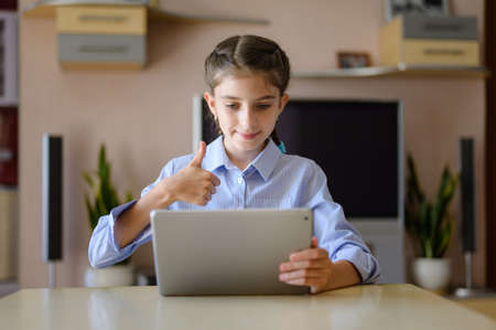 Joyful girl sitting at a table with a tablet. Girl communicates via the Internet and shows thumb up.の写真素材