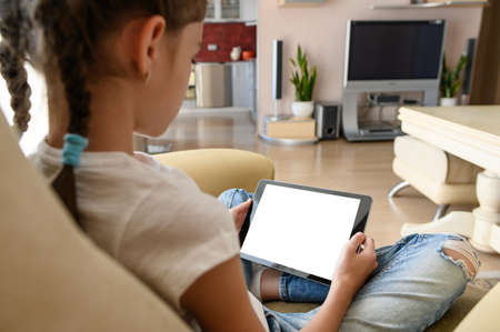 A girl sits at home on an armchair and holds in her hands a tablet with a white blank display. Use modern gadgets in your home everyday environment.の写真素材