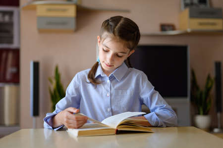 A girl sits at a table and carefully reads a book. Getting ready to turn the page.の写真素材