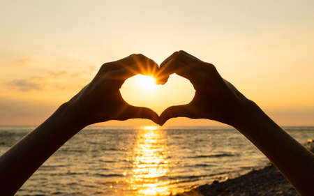 Human hands show the shape of a heart at sunset against the background of the sea. Sun rays at...