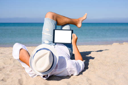 A man lies on a sandy beach with a tablet. The sea is in the background. Empty space for text. A man in a white shirt and denim shorts.の写真素材