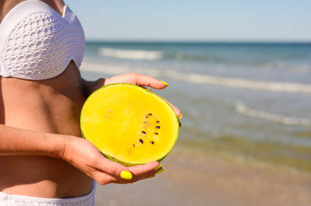 The girl on the background of the sea holds a yellow watermelon. Delicious fruits on the beachの写真素材