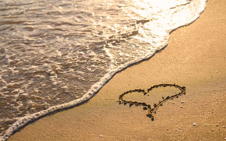 A heart is drawn on the sandy beach. The sea wave washes the beach at sunset.の写真素材