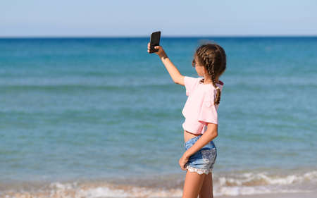 A girl stands on the beach near the sea and takes a selfie. A modern girl takes pictures of herself...
