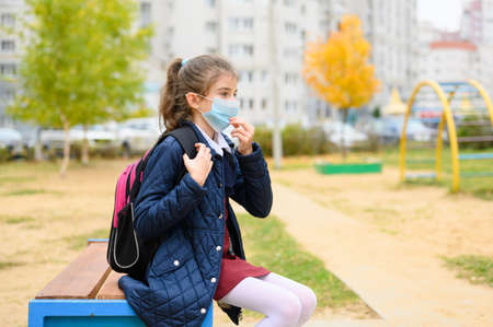 A schoolgirl adjusts a medical mask on her face. The girl sits on the street wearing a medical mask.の写真素材