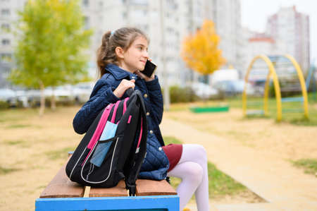 A schoolbag with a medical mask stands on a bench. In the background, a schoolgirl is talking on a cell phone. Focus on mask and briefcase.の写真素材