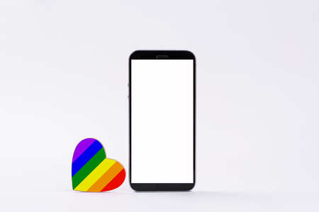 A small heart painted in the color of the LGBT flag stands beside a smartphone with a blank white display. Items on a white background. The concept of LGBT relationships and declaration of love with through messages.の写真素材