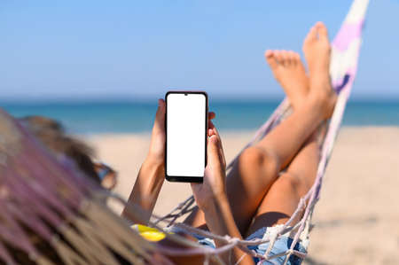 Smartphone with a blank white display in the hands of a girl against the background of the sea. The girl lies on a hammock on the beach with a phone in her hands.の写真素材