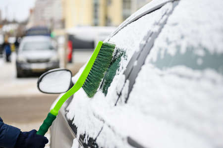 The process of cleaning snow from a car with a brush. A green brush cleans snow from the side window of a car in a parking lot. Focusing on the brush.の写真素材