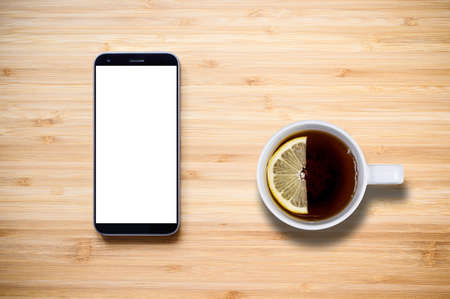 A cup of black tea with lemon on a wooden table next to a mobile phone. Phone with blank white display. View from above.の写真素材