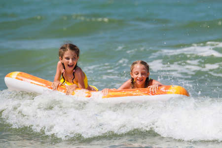 Two joyful girls ride on a water mattress on the sea waves. The sisters are swimming in the sea.の写真素材