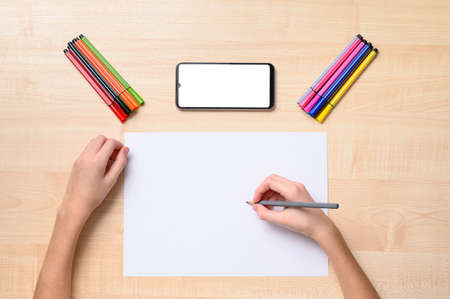A white sheet, pencils, a phone with a blank white display and lie on the table. The hand lies on the sheet and holds a pencil. Online lesson conceptの写真素材