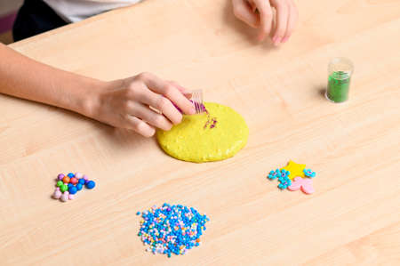 The girl prepares slime at home. Girl sprinkles yellow slime with pink sparkles. Focus on hand and jar of glitterの写真素材