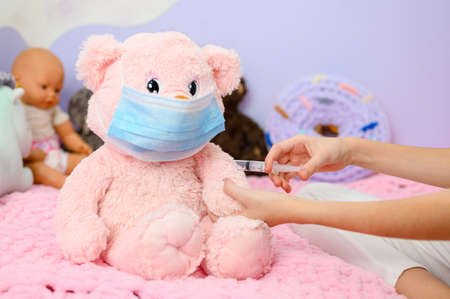 A girl is vaccinated against the coronavirus to her teddy bear, who is blown away in a medical mask. Focusing on the syringe.の写真素材