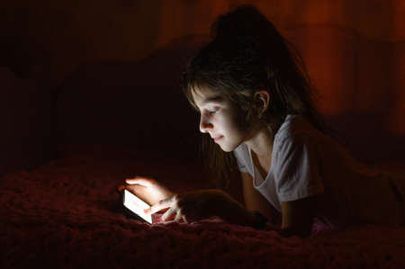 The girl lies on the bed with a phone in her hands. The girl plays on the phone late in the evening. Side view.の写真素材