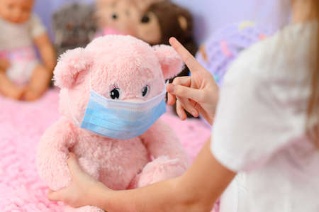 The girl plays with dolls at home. The girl shows her hand to the teddy bear about the importance of wearing a medical mask during coronavirus. Focus on the girl's hand.の写真素材