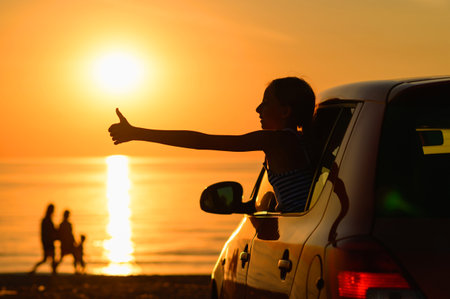 The girl enjoys a trip to the sea by car. In the background there is a beautiful sunset and the silhouette of a family walking along the seashore. Auto travel concept.の写真素材