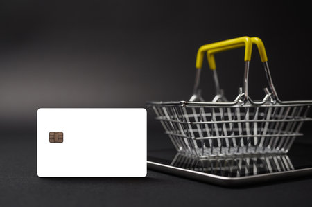 Online shopping concept. Blank white form of a bank card on a dark background. Nearby, the shopping basket is on the tablet screen.の写真素材