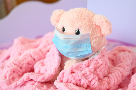 A pink teddy bear in a medical mask and a thermometer sits covered with a pink blanket. Coronovirus or flu disease concept. Focusing on the bear's eye.の写真素材