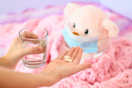 Pills in children's hands and a glass of water for a sick toy bear. The child plays in his room. Virus treatment concept. Focus on pills.の写真素材