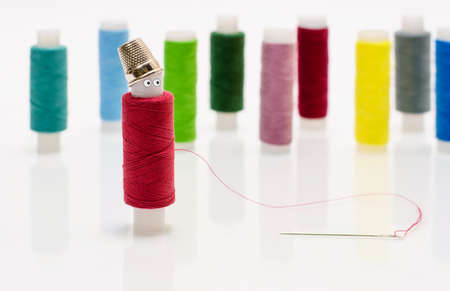 A red spool of thread for sewing with funny eyes and a thimble on his head against the background of multi-colored spools of thread. Focusing on the red coil. Sewing lovers concept.の写真素材