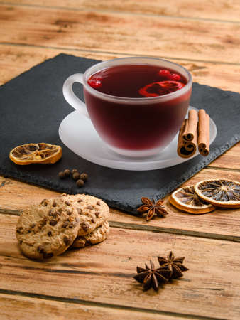 Vertical composition of a cup of hot fruit tea or mulled wine on an old wooden table. Next to it is a cup of biscuits, cinnamon and dry lemon rings. Focusing on the drink cup.の写真素材