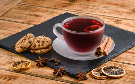 A cup of hot fruit tea or mulled wine on a slate board. Next to it is a cup of biscuits, cinnamon and dry lemon rings.の写真素材