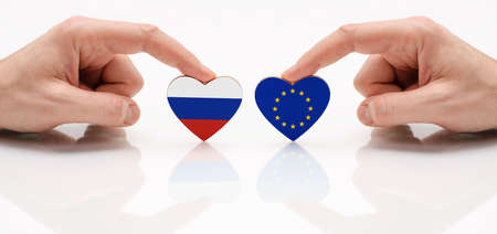 The concept of friendship and diplomatic relations between Russia and the European Union. Two male hands are holding flags in the shape of a heart on a glass table. The flag of Russia and the European Union as a symbol of cooperation and respect.の写真素材
