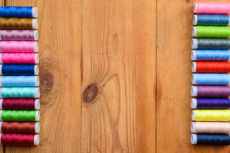 Lots of multicolored spools of thread for sewing are stacked evenly around the edges on an old wooden table. Empty white space in the center of the frameの写真素材