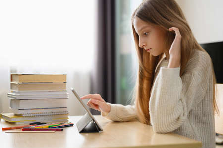 A schoolgirl with long blond hair uses a tablet at her desk. A stack of textbooks in the background. Home distance learning concept.の写真素材