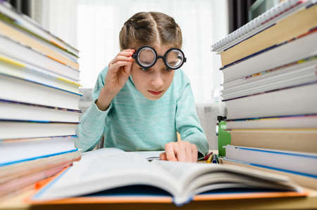 A girl in funny round glasses with large lenses looks at a book in surprise. There are many textbooks nearby.の写真素材