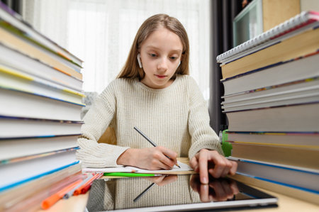 A modern girl is doing her homework or preparing for an exam using a tablet and the Internet. The girl looks carefully at the display of the tablet. A bunch of books and textbooks nearbyの写真素材