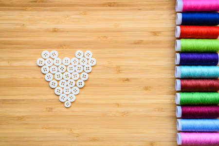The spools of sewing thread are laid flat on the wooden table next to the white buttons. Buttons are laid in the shape of a heart.の写真素材