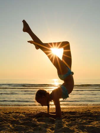 The sun's rays shine through the legs of a gymnast on the beach. The girl performs a handstand and holds her legs in the shape of a triangle. Sunset at sea.の写真素材