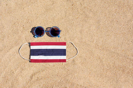 A medical mask in the color of the Thailand flag lies on the sandy beach next to the glasses. The concept of organizing a safe beach holiday in Thailand during the coronavirus pandemic.の写真素材