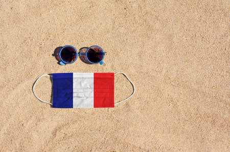 A medical mask in the color of the French flag lies on the sandy beach next to the glasses. The concept of organizing a safe beach holiday in France during the coronavirus pandemic.の写真素材