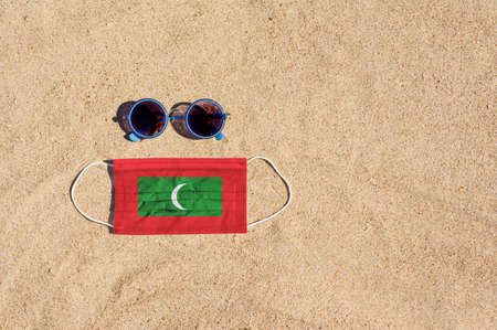 A medical mask in the color of the Maldives flag lies on the sandy beach next to the glasses. The concept of organizing a safe beach holiday in Maldives during the coronavirus pandemic.の写真素材