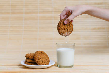 Children's hand dries oatmeal cookies in a glass of milk. Cookies lie on a white saucer on a wooden background.の写真素材