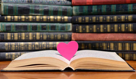 A pink heart lies between the pages in an open book. Yea in the background are piles of old books.の写真素材