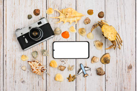 Seashells, a crab, an old camera and an airplane figurine on a wooden table. In the center is a smartphone with a blank white display. Travel and tourism concept.の写真素材