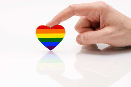 Gay Pride Flag in the shape of a heart. A man's hand holds a heart in the shape of the LGBT flag on a white glass surface. The concept of tolerance and equality for LGBT peopleの写真素材