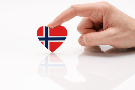 Love and respect Norway. A man's hand holds a heart in the shape of the Norway flag on a white glass surface. The concept of Norwegian patriotism and pride.の写真素材