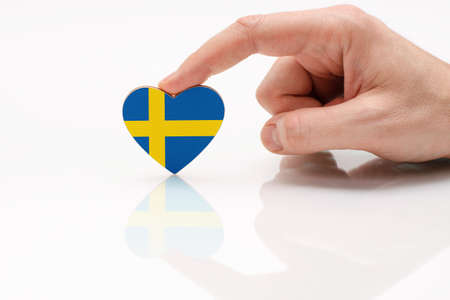 Love and respect Sweden. A man's hand holds a heart in the shape of the Sweden flag on a white glass surface. The concept of Swedish patriotism and pride.の写真素材