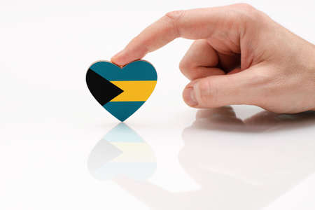 Flag of the Bahamas. Love and respect Bahamas. A man's hand holds a heart in the shape of the Bahamas flag on a white glass surface. The concept of patriotism and tourismの写真素材