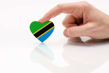 Tanzania flag. Love and respect Tanzania. A man's hand holds a heart in the shape of the Tanzania flag on a white glass surface. The concept of patriotism and tourism.の写真素材